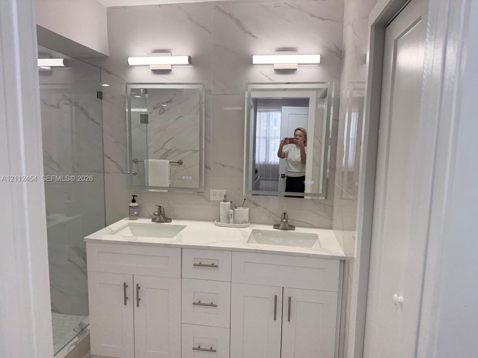 Double vanity featuring white shaker-style cabinetry, white stone-composite countertops, integrated rectangular sinks, and illuminated wall-mounted mirrors with large-format tile surround