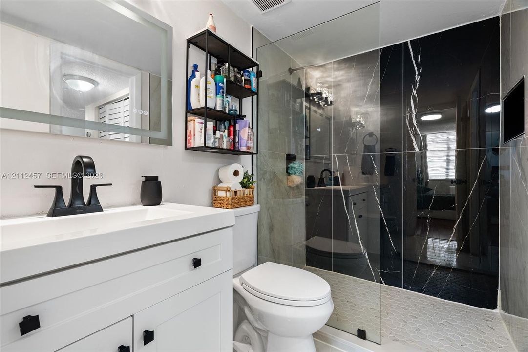 White vanity with matte black hardware, integrated sink, matte black widespread faucet, LED-backlit mirror, and walk-in shower with floor-to-ceiling dark stone-patterned tile and frameless glass partition