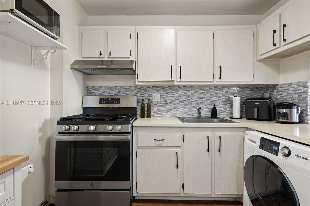 Kitchen featuring white cabinetry with matte black hardware, stainless steel gas range, stainless steel range hood, stone-mosaic tile backsplash, and integrated laundry appliance