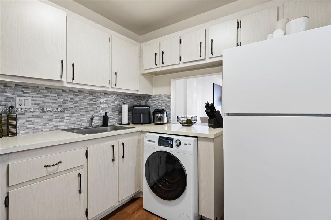 Kitchen featuring light wood-finish cabinetry, a multi-tone linear tile backsplash, solid-surface countertops, and an integrated washer-dryer unit