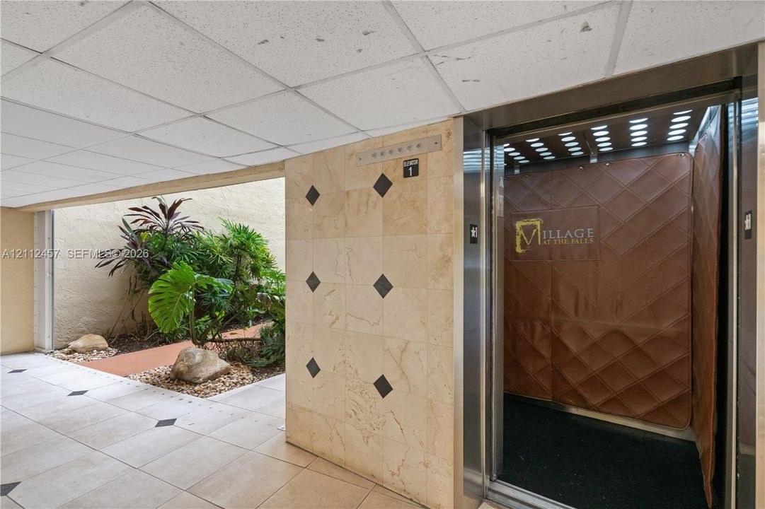 Elevator lobby featuring square tile flooring with black accent insets, polished light-toned stone wall cladding, and an integrated plant bed with stone ground cover