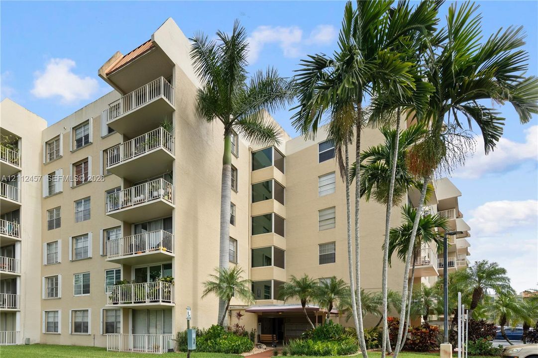 Multi-story building exterior featuring stucco cladding, stacked private balconies with metal railings, and mature palm tree landscaping