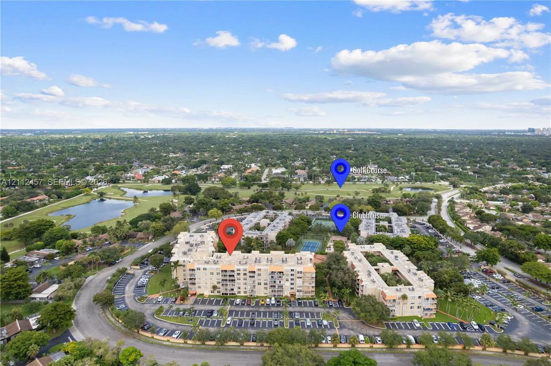 Aerial perspective of a multi-story residential complex featuring paved surface parking, tennis courts, and proximity to green space and water features