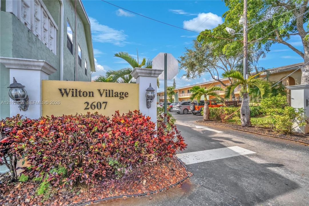 Community entrance featuring stone-pillar signage, wall-mounted coach lighting, paved access road, and mature tropical landscaping