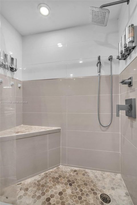 Walk-in shower featuring large-format beige wall tile, contrasting white upper wall tile, hexagonal stone floor tiling, a built-in corner bench, and a dual-head rainfall and handheld shower system