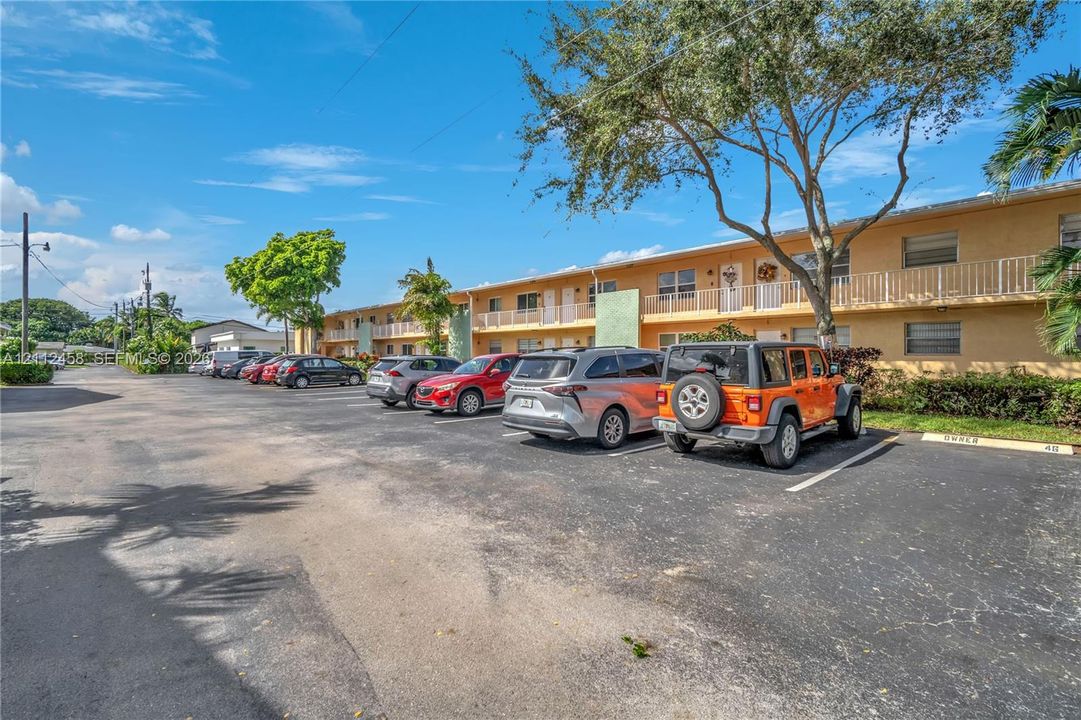 Two-story building exterior featuring stucco siding, metal-railing balconies, asphalt parking lot, and mature landscaping
