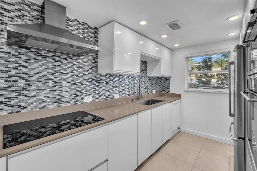 Kitchen featuring flat-panel white cabinetry, solid-surface countertops, a stainless steel range hood, built-in cooktop, and a glass-tile backsplash