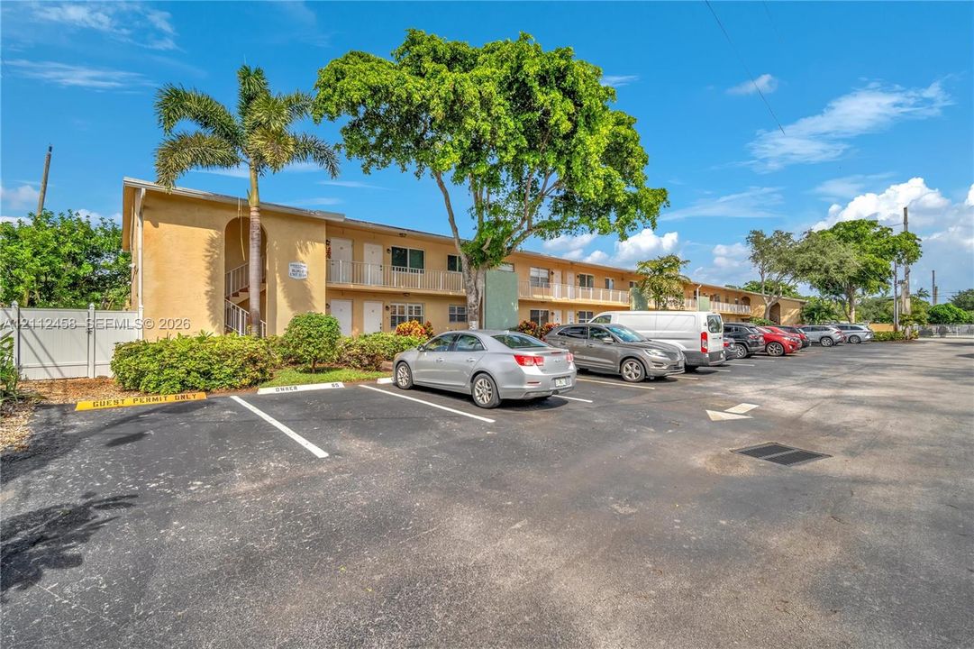 Multi-story exterior featuring a stucco facade, covered balconies, paved surface parking, and mature palm and shade trees