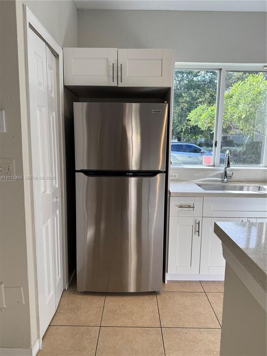 Stainless steel refrigerator with overhead built-in cabinetry, shaker-style white lower cabinets, stone-composite countertops, and tiled flooring