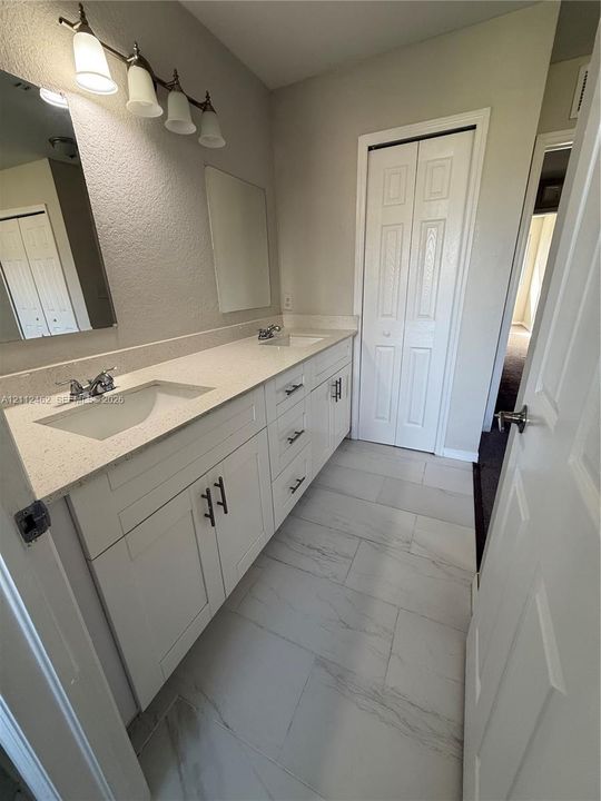 Dual vanity featuring a light-toned stone-composite countertop, white shaker-style cabinetry with matte black hardware, integrated rectangular basins, and large format rectified tile flooring
