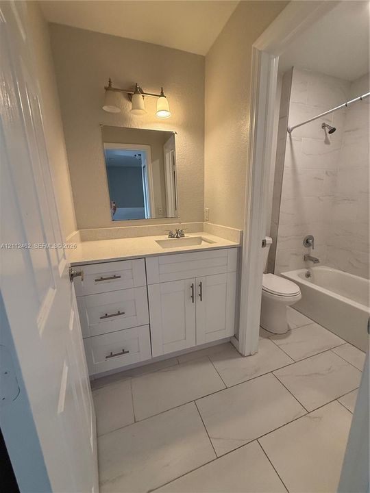 White vanity with stone-look countertop, integrated rectangular sink, three-light wall-mounted fixture, and large-format tile flooring