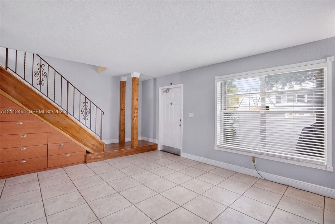 Interior featuring tile flooring, a large window with horizontal blinds, built-in wooden under-stair storage, decorative wrought-iron railing, and dual wooden support columns