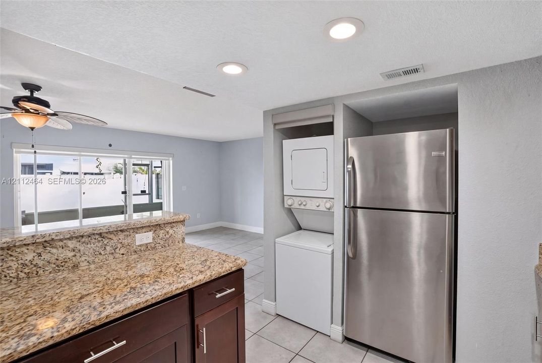 Kitchen area featuring stone-finish countertops, dark wood-finish cabinetry, stainless steel refrigerator, stacked laundry unit, and ceramic tile flooring