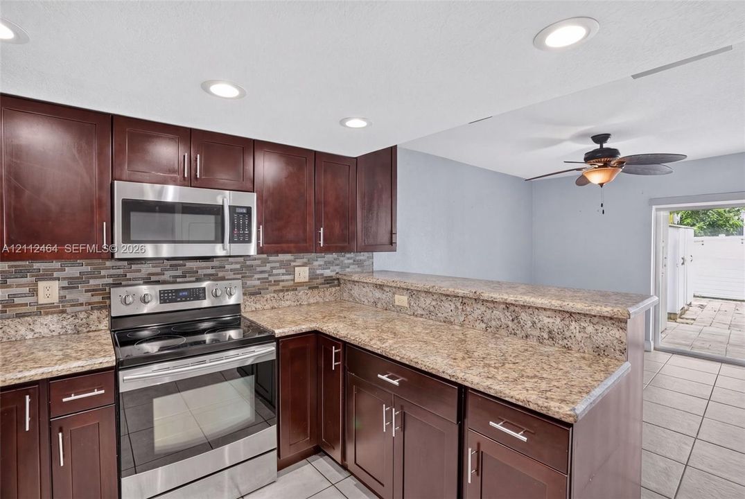 Kitchen featuring dark wood cabinetry, stainless steel appliances, stone-patterned countertops, a multi-tone tile backsplash, and recessed lighting