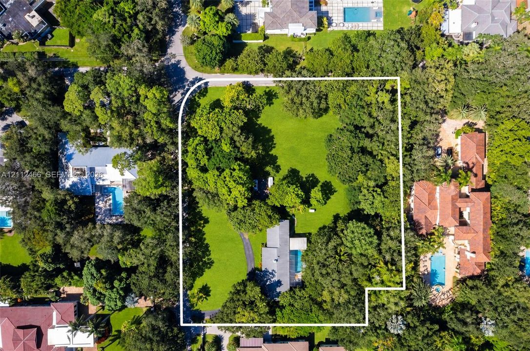 Aerial perspective of a sprawling corner lot featuring mature trees, an expansive lawn, a paved driveway, and an inground swimming pool