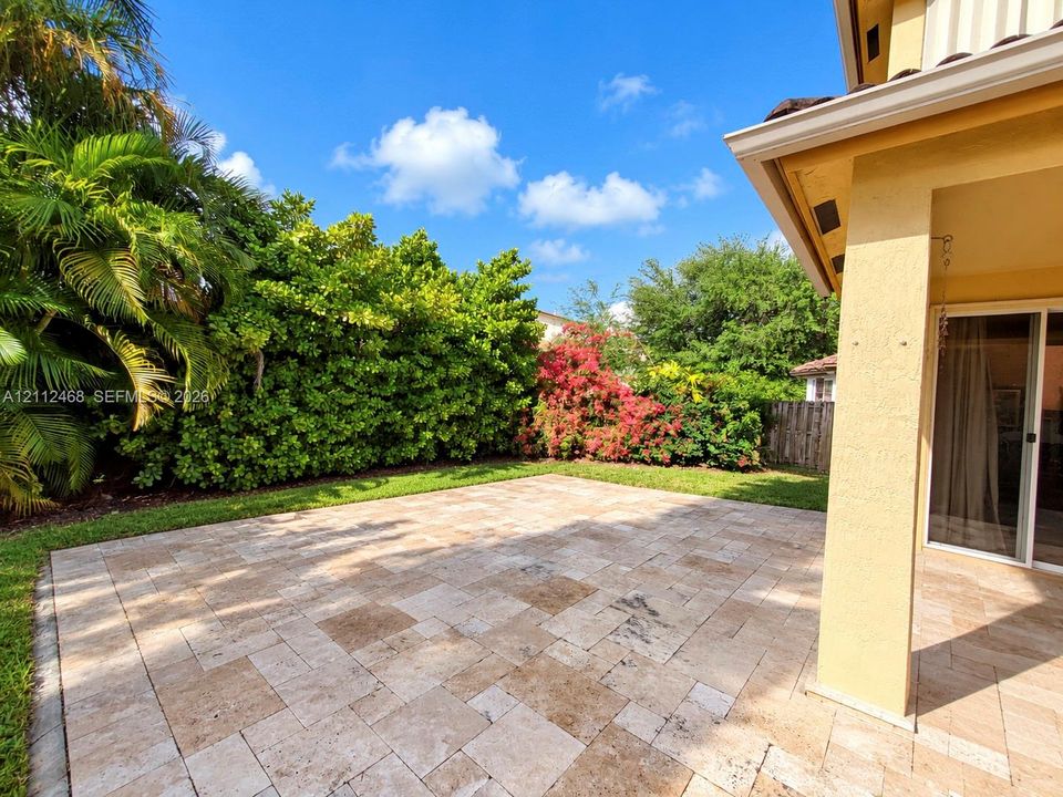 Covered patio featuring stone-tiled flooring, landscaped lawn, mature privacy hedges, and flowering shrubs