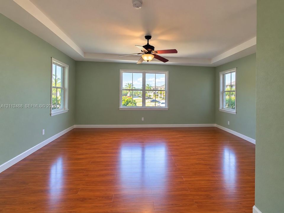 Room featuring wood-finish flooring, a tray ceiling, and a ceiling fan with integrated light fixture, illuminated by three double-hung windows