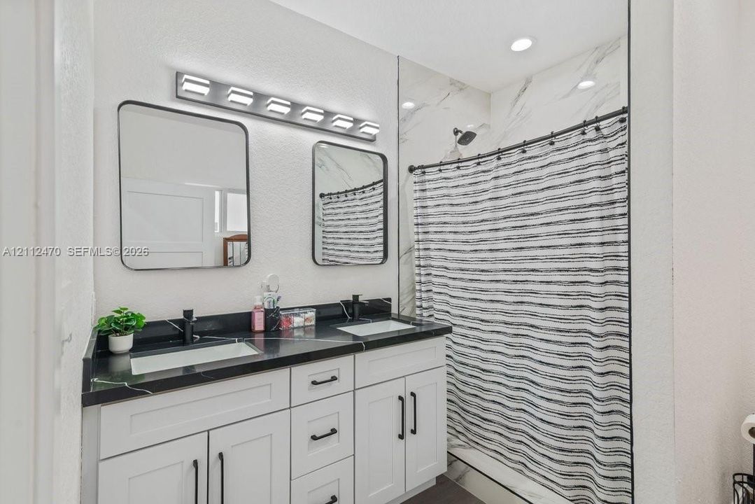 Dual-sink vanity with white shaker-style cabinetry, dark stone-finish countertop, dual rectangular undermount sinks, and matte black hardware, accompanied by a shower surround featuring large-format wall tile