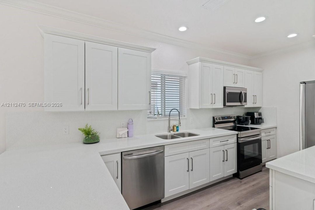 Kitchen featuring white shaker-style cabinetry, solid-surface countertops, recessed lighting, and stainless steel appliances, including an over-the-range microwave and integrated dishwasher, complemented by wood-finish flooring