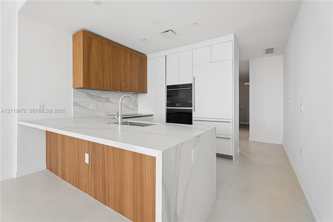 Kitchen featuring a waterfall-edge island with stone-finish countertop, wood-finish cabinetry, built-in wall oven and microwave, panel-ready refrigerator, and stone-finish backsplash