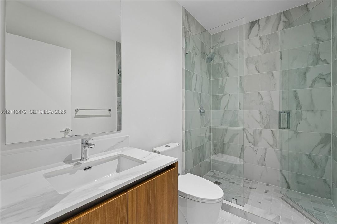 Bathroom featuring a wood-finish vanity with a stone-top integrated sink, a large wall-mounted mirror, a walk-in shower with floor-to-ceiling tile, and a glass partition