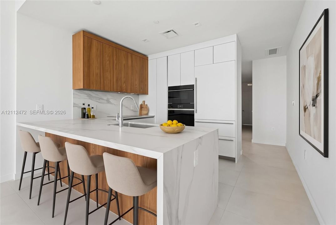 Kitchen featuring waterfall-edge stone island with integrated sink, flat-panel wood cabinetry, white floor-to-ceiling cabinetry, stone backsplash, and built-in wall oven. VIRTUALLY STAGED