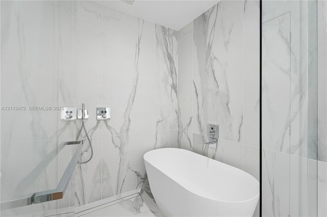 Freestanding white soaking tub with wall-mounted chrome fixtures, floor-to-ceiling large-format stone-finish tile surround, and built-in linear floor drain
