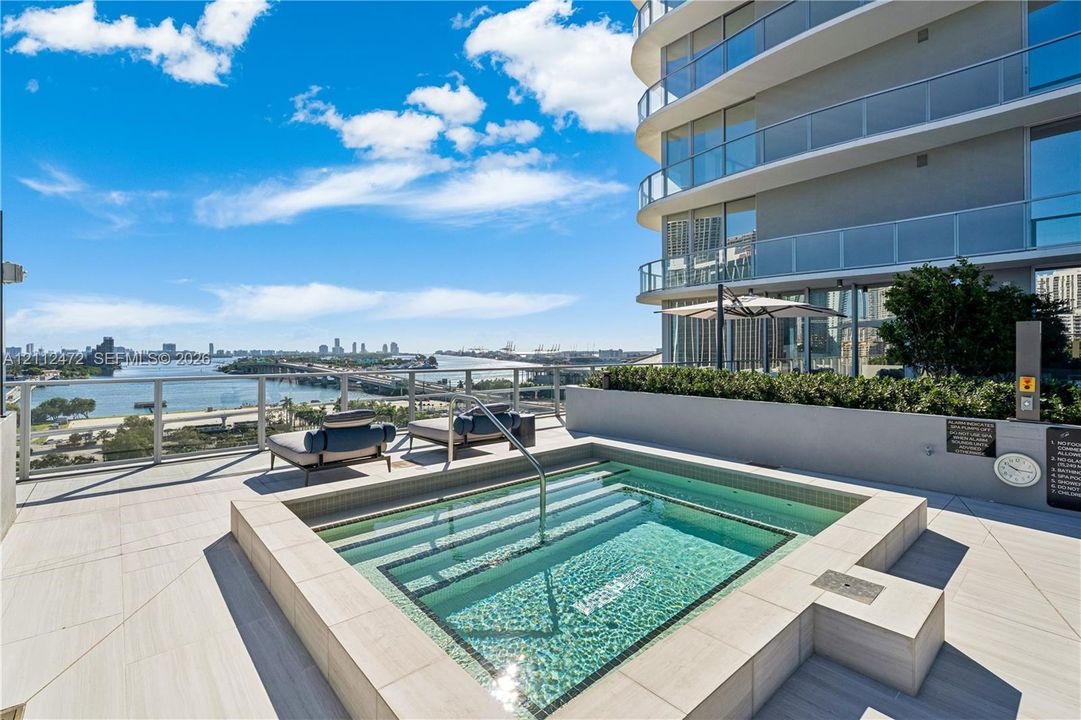 Terrace featuring a recessed square spa, large-format tile flooring, a glass panel railing, and a multi-story exterior with curved balconies