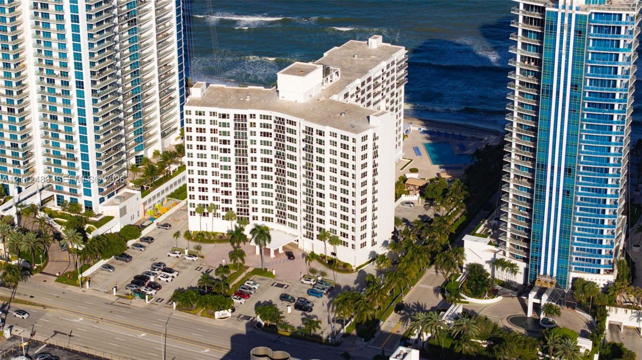 Aerial perspective of high-rise building featuring proximity to the coastline, paved surface parking, mature palm trees, and an outdoor swimming pool