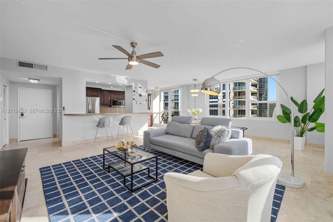 Open-concept living space featuring natural stone flooring, large windows, dark wood-finish cabinetry, a breakfast bar, and a ceiling fan with integrated lighting