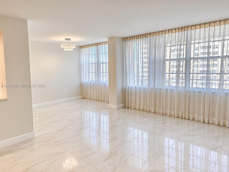 Large-format neutral stone-finish tile flooring, expansive windows with wall-to-wall drapery, and a tiered crystal-style flush-mount ceiling fixture