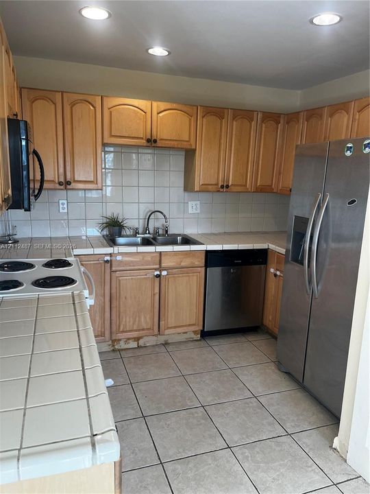 Kitchen featuring light wood cabinetry, tiled countertops and backsplash, stainless steel refrigerator and dishwasher, and ceramic tile flooring