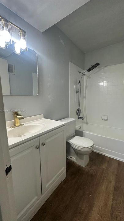 Bathroom featuring a white vanity cabinet, wood-finish flooring, gold-toned faucet, rectangular mirror with rounded corners, and a white shower-tub combination