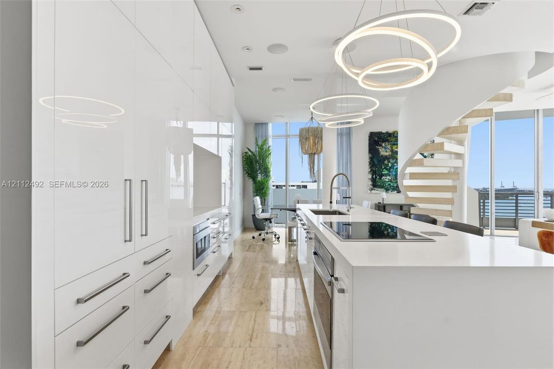 High-gloss cabinetry, stone-surfaced kitchen island, integrated stainless steel appliances, circular pendant lighting, and a sculptural spiral staircase with floating treads