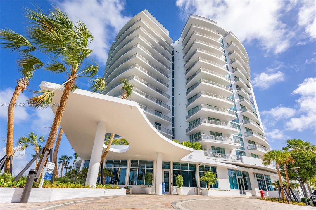 High-rise residential building featuring a sculpted architectural porte-cochère, stacked private balconies, floor-to-ceiling windows, and paved motor court