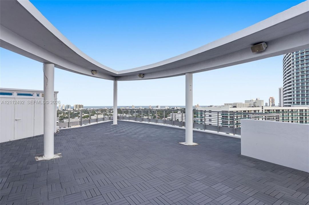 Rooftop terrace featuring dark modular interlocking flooring, a curved architectural canopy supported by cylindrical columns, and metal perimeter railing