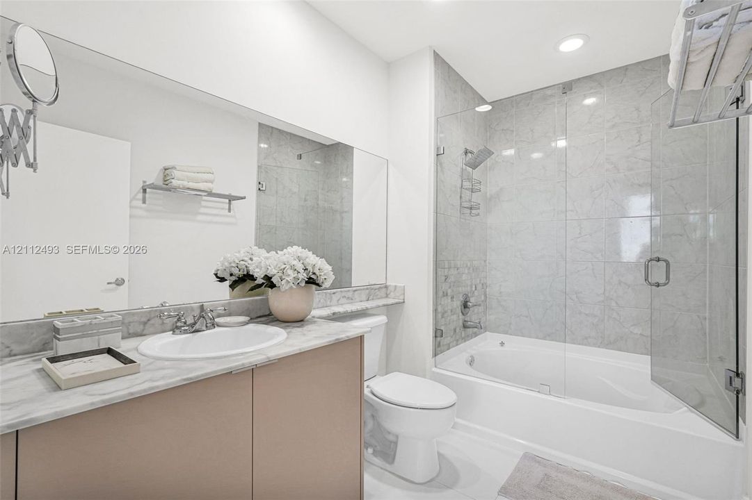 Bathroom featuring a vanity with a light stone countertop, an integrated sink, a large wall-mounted mirror, a tiled shower-tub combination with a glass enclosure, and recessed lighting