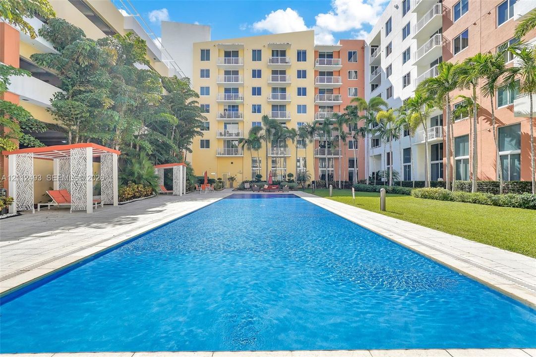 Rectangular swimming pool surrounded by stone paver decking, manicured lawn, architectural cabanas, and mature palm trees