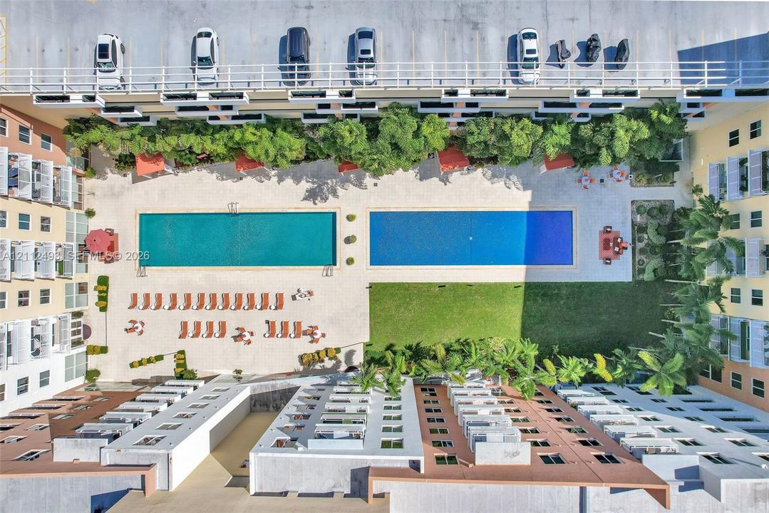 Aerial view of a complex featuring two rectangular swimming pools, expansive paved pool deck, manicured lawn, and perimeter landscaping with mature palm trees