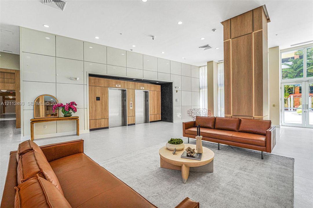 Lobby featuring panelized wall finishes, double elevator bank with wood-tone accents, recessed lighting, expansive glass entryway, and large-format flooring