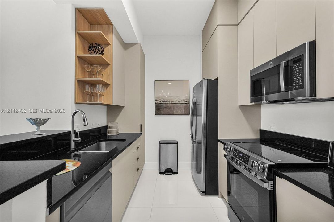 Kitchen featuring flat-panel cabinetry with light-tone finish, black stone countertops, open wood-finish shelving, stainless steel appliances, and large-format tile flooring