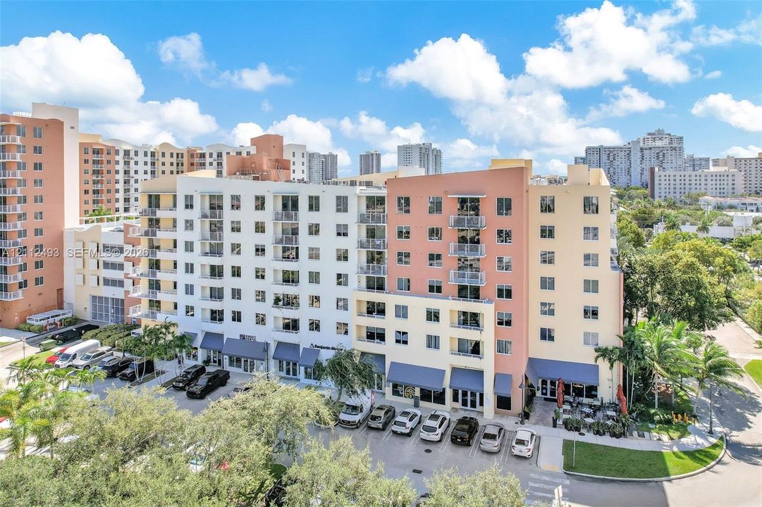 Multi-story exterior featuring a facade with integrated balconies, ground-level storefronts with canvas awnings, and an asphalt parking area