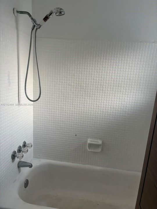 Bathtub with white square tile surround, wall-mounted hand-held shower head, integrated soap dish, and three-handle chrome faucet fixture