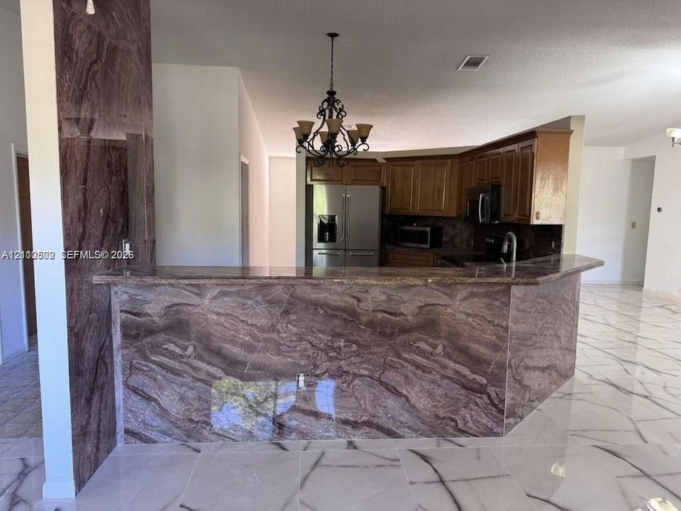 Kitchen featuring large-format tile flooring with veining, stone-clad breakfast bar, wood cabinetry, stainless steel appliances, and ornamental wrought iron chandelier