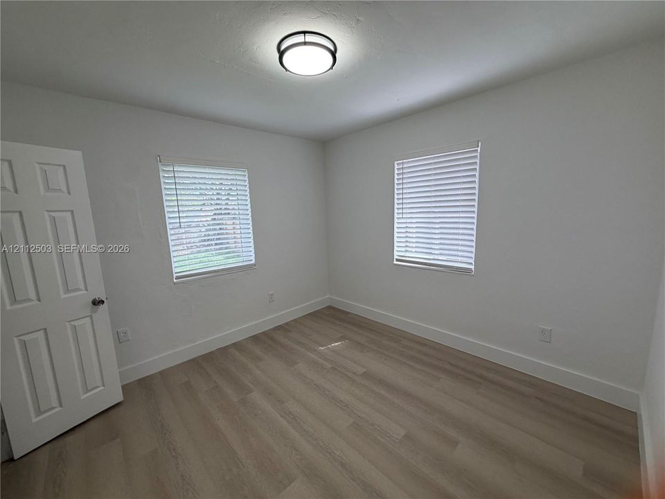 Neutral-toned wood-finish flooring, two windows with installed horizontal blinds, solid-core four-panel interior door, and modern circular flush-mount ceiling lighting
