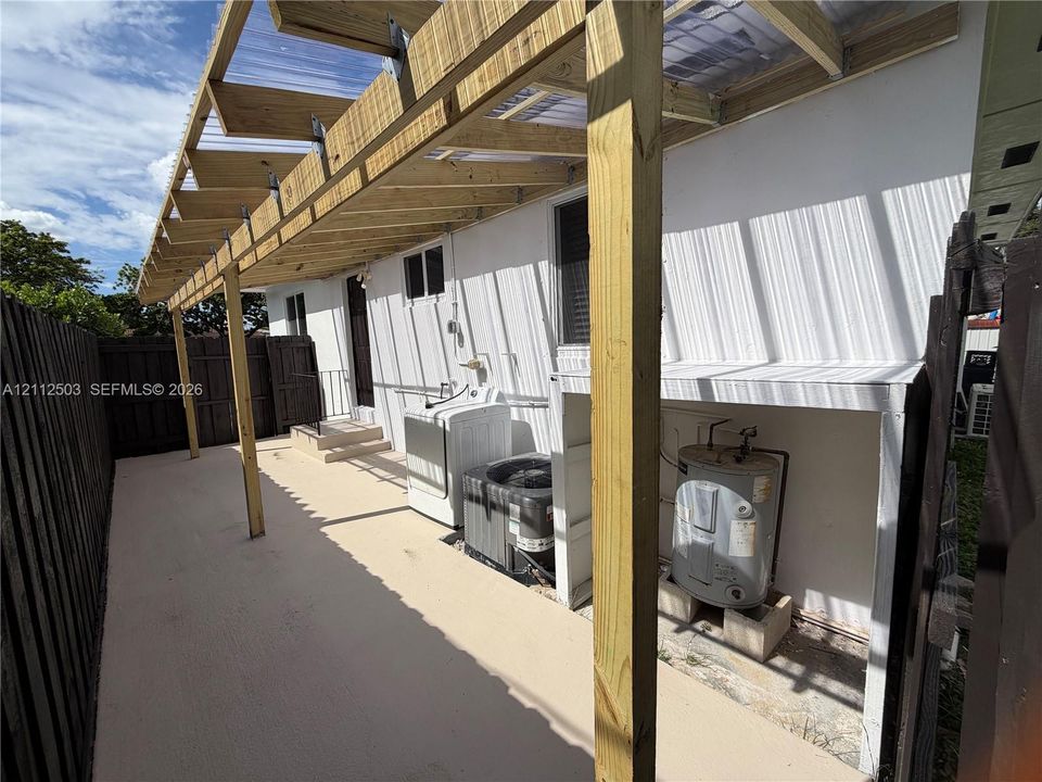 Covered exterior patio featuring a wood-frame structure with translucent roofing, concrete flooring, and an enclosed wood privacy fence