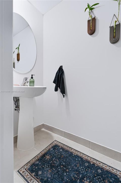 Pedestal sink with a single-handle chrome faucet, frameless circular wall mirror, and neutral tile flooring with coordinated tile baseboards