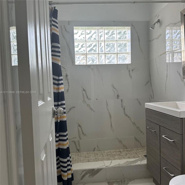 Bathroom featuring large-format wall tiles with veined detailing, glass block window, stone-look pebble tile shower floor, and dark wood-finish vanity with a rectangular integrated sink