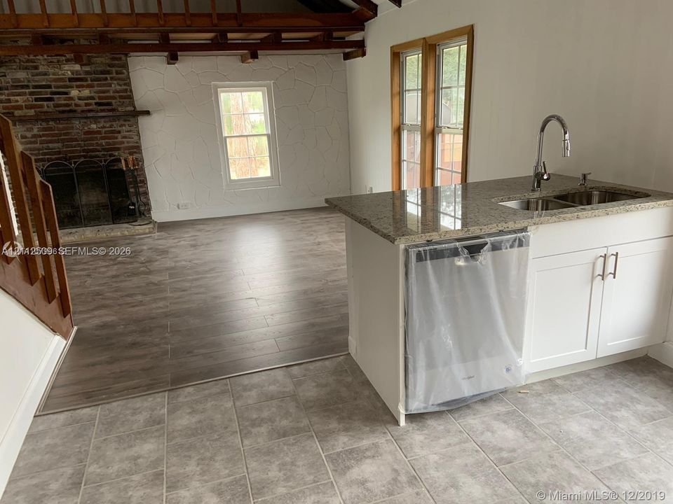 Kitchen island featuring stone countertops, integrated stainless steel sink, and white cabinetry, paired with wood-finish flooring, a full-height brick fireplace, and exposed structural ceiling beams