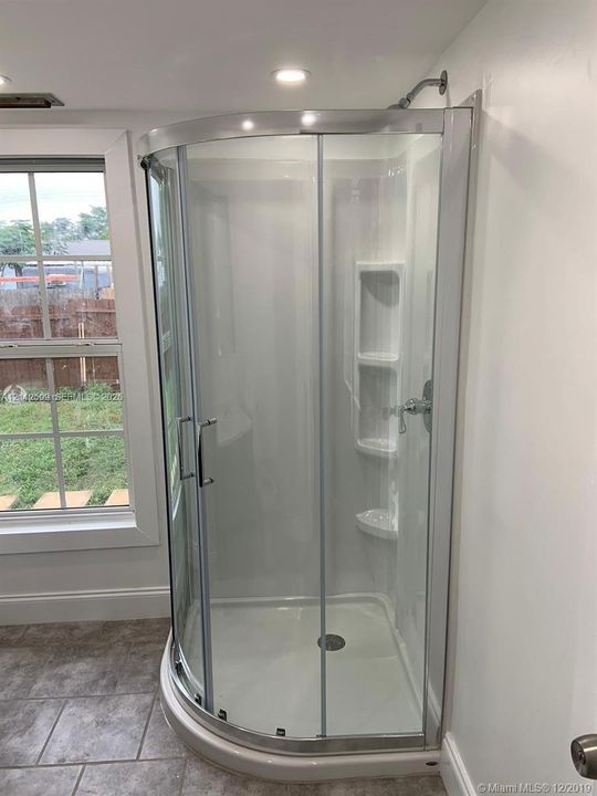 Corner shower stall featuring a curved sliding glass door, white surround with built-in shelving, chrome-finish hardware, and tile flooring